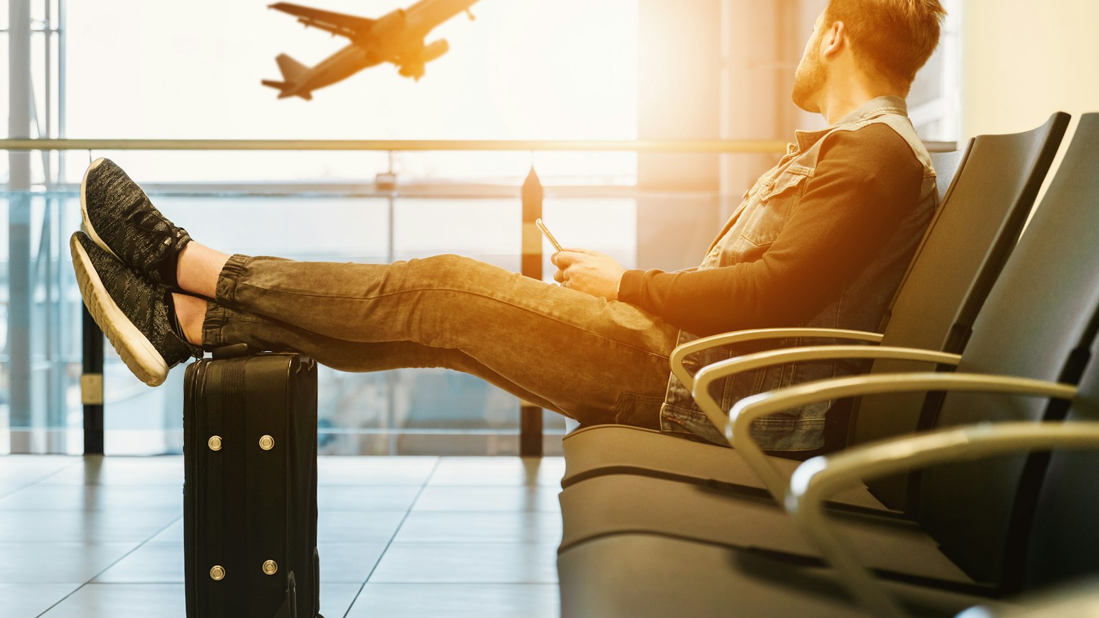Viajero espera en la sala del aeropuerto con los pies sobre su maleta mientras despega un avión